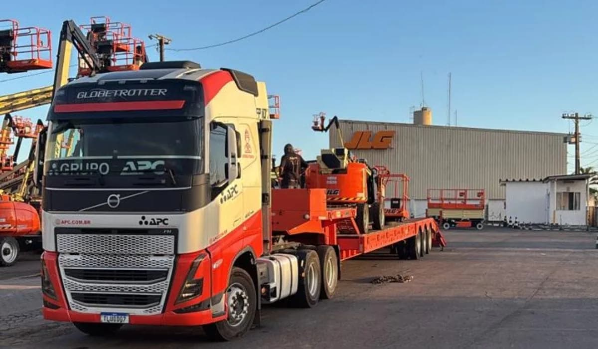 Carreta do Grupo APC carregando uma plataforma de lança JLG no pátio da empresa, com a frota ao fundo